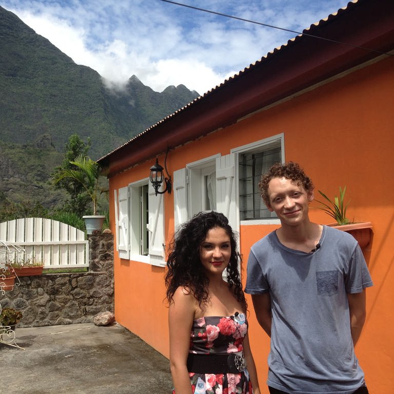 Eine junge Frau und ein Mann vor einem orangefarbenen Haus; im Hintergrund ein Berg.