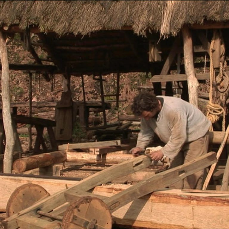 Ein Mann bearbeitet einen kleinen Wagen aus Holz mit einem Ziehmesser.