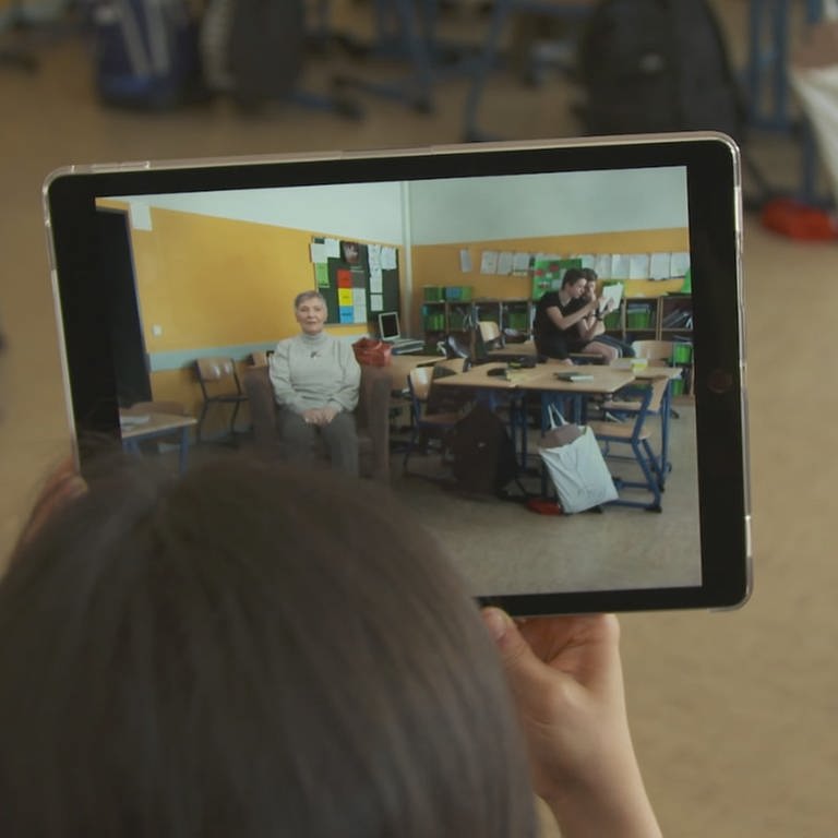 Auf einem Tablet sieht eine Frau einen Klassenraum und die Holografie einer Zeitzeugin