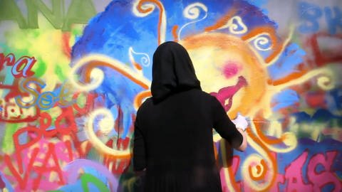 Frau mit schwarzem Kopftuch beim Sprühen eines Graffito.