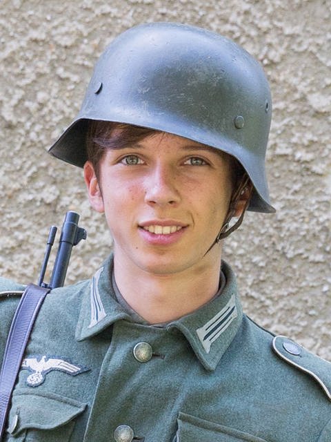 Ein Jugendlicher in Uniform und Soldatenhelm. Auf der Uniform ist ein Zeichen der NSDAP gestickt.