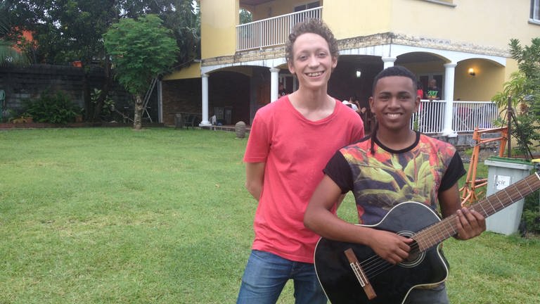 Zwei junge Männer stehen vor einem Haus. Einer hält eine Gitarre.