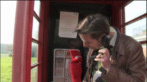 Ein Mann und eine rote Strumpfhandpuppe telefonieren in einer Telefonzelle.