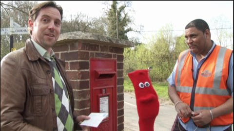 Ein Mann steht vor einem Briefkasten. Neben ihm eine rote Strumpfhandpuppe und ein Postbote.