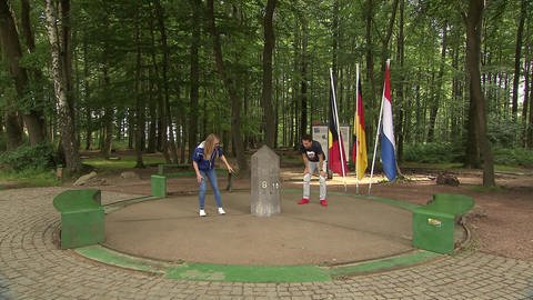 Ein Mann und eine Frau stehen im Wald um einen kleinen Steinobelisk. Im Hintergrund wehen die Fahnen von Deutschland, Belgien und den Niederlanden.
