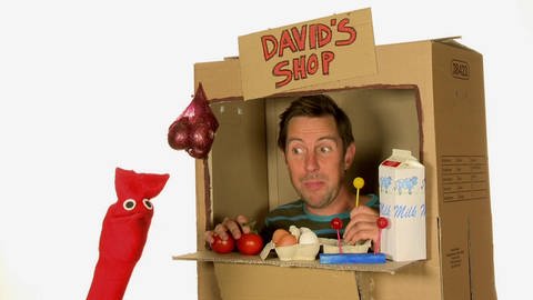 Ein Mann sitzt in einem Einkaufsladen aus Pappe. Auf einem Schild steht „David‘s Shop“. 