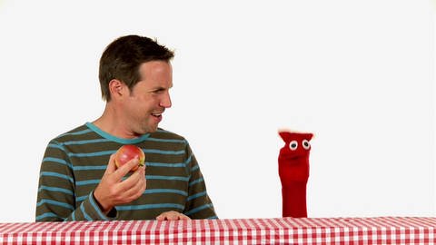 Ein Mann hält einen Apfel in der Hand und schaut dabei bestürzt eine rote Strumpfhandpuppe an. 
