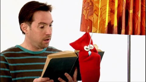 Ein Mann liest ein Buch, eine rote Strumpfhandpuppe macht eine Stehlampe neben ihm aus.