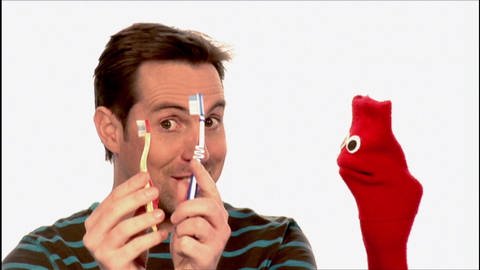 Ein Mann hält zwei Zahnbürsten hoch. Neben ihm eine rote Strumpfhandpuppe.