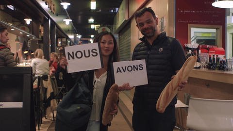 Eine Frau und ein Mann stehen in einem Restaurant. Sie halten Schilder hoch mit "Nous" und "avons", der Mann hält ein Baguette.