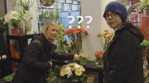 Ein Junge und eine Verkäuferin stehen in einem Blumenladen, neben seinem Kopf schweben drei Fragezeichen.