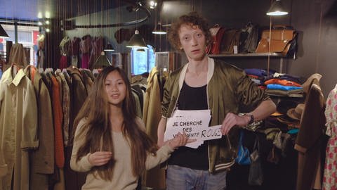 Ein junger Mann und eine junge Frau stehen in einem Kleiderladen. Der Mann hält ein Schild mit der Aufschrift "Je cherche des vêtements rouge".
