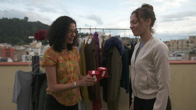 Zwei Frauen stehen auf einer Terasse vor einer Kleiderstange. Die eine Frau hält der anderen zwei rote Würfel entgegen.