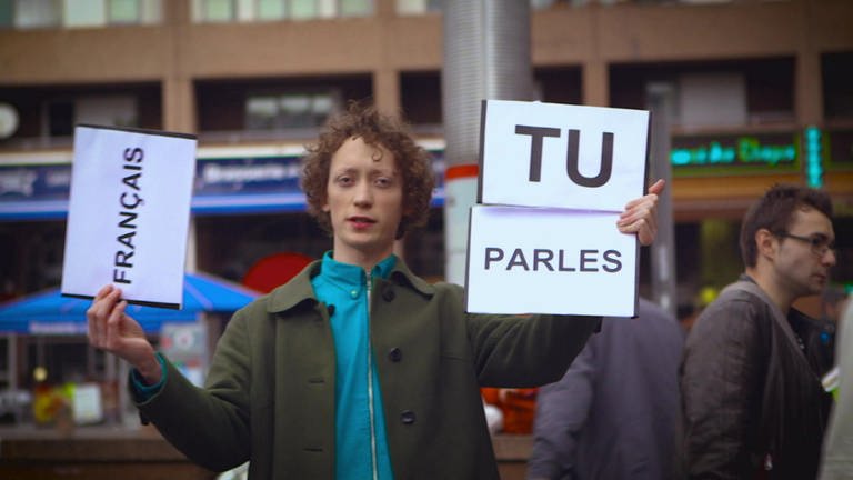 Ein junger Mann steht auf der Straße und hält Schilder hoch, darauf steht "Tu parles français"