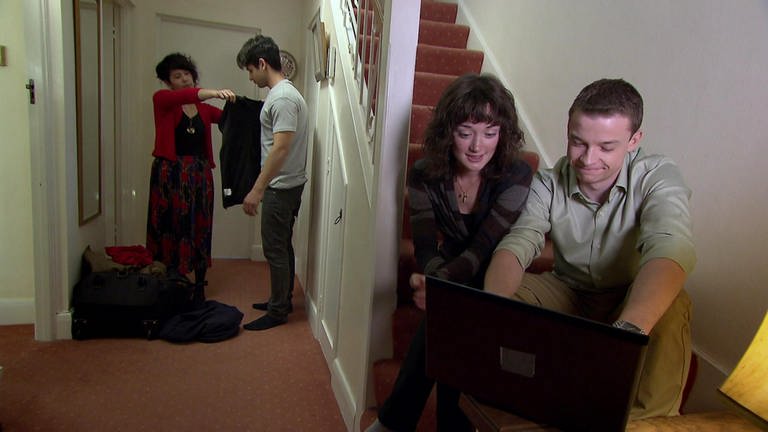 Ein junger Mann und eine junge Frau sitzen auf einer Treppe und schauen auf einen Computer. Neben der Treppe probieren zwei andere junge Erwachsene Kleidung an.