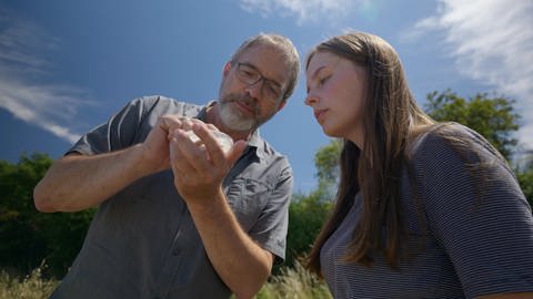 Zwei Forscher stehen neben einem Feld auf einem Blühstreifen und untersuchen eine Probe 