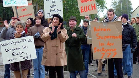 Frauen und Männer, die meisten winterlich angezogen, stehen auf einem Platz, klatschen und halten selbst gemalte Protesttafeln hoch. Einige der Aufschriften: „Wehrt Euch“, „Eugen Forever“, „Sackgasse 777. Wir sind bei Euch, jeden Tag!“