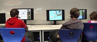 Drei Kinder sitzen mit dem Rücken zur Kamera vor Computern und spielen das Lernspiel Deutsch lernen mit Mumbro und Zinell. 