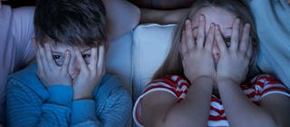Zwei Kinder sitzen im Schein eines Fernsehers und gucken durch die Hände, mit denen sie sich die Augen halb zuhalten