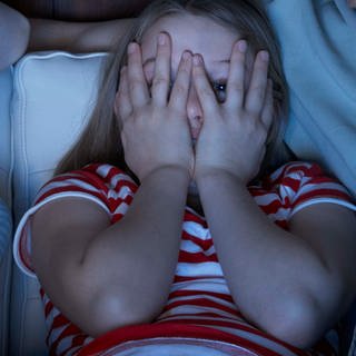 Zwei Kinder sitzen im Schein eines Fernsehers und gucken durch die Hände, mit denen sie sich die Augen halb zuhalten