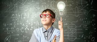 Junge mit Brille steht vor einer beschriebenen Tafel, neben ihm schwebt eine helle Glühbirne