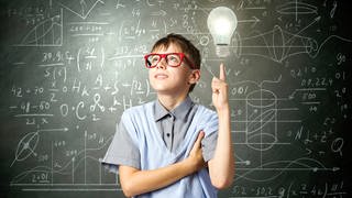 Junge mit Brille steht vor einer beschriebenen Tafel, neben ihm schwebt eine helle Glühbirne