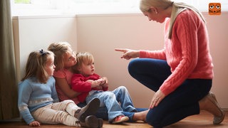 Eine Frau zeigt ermahnend mit dem Zeigefinger auf ein paar Kinder, die ihr gespannt zuhören.