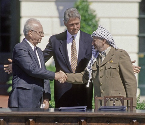 Yitzhak Rabin und Jassir Arafat, geben sich die Hand. Bill Clinton breitet die Arme über ihnen aus.