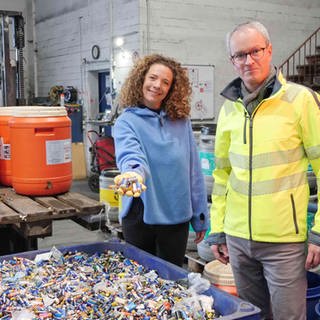 neuneinhalb-Reporterin Gesa und Florian Clever stehen vor einer großen blauen Plastikwanne voller Altbatterien in einer Recycling-Anlage. Im Hintergrund stehen bunte Plastikfässer voller Batterien. Gesa hält eine Handvoll Batterien in Richtung Kamera.
