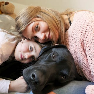 neuneinhalb-Reporterin Jana und Siona kuscheln mit Sionas schwarzem Labrador Pluto.