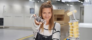 neuneinhalb-Reporterin Jana hält einen Bionik-Roboter in der Hand, der einer Ameise nachempfunden ist. Auf dem Tisch vor ihr steht ein Roboterarm, der aussieht, wie ein Elefantenrüssel.