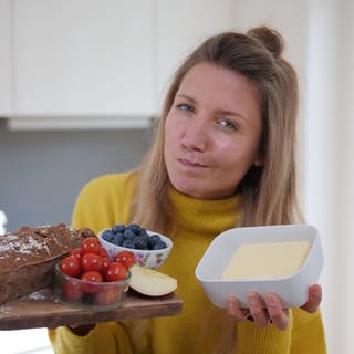 Jana schaut kritisch in die Kamera. Sie hält unterschiedliche Lebensmittel ins Bild, darunter Butter, Tomaten, Heidelbeeren, ein Brot und einen Apfel.