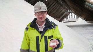 Robert steht in einer Halle, in der Salz aufgetürmt ist. Er trägt Schutzkleidung und hält Salz in der Hand.