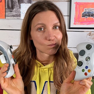 Reporterin Jana sitzt auf einer Couch. Sie hält zwei Controller in den Händen und schaut fragend in die Kamera.