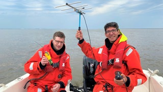 Reporter Robert und der Unterwasserlärm-Forscher Joseph Schnitzler sitzen in einem Boot. Robert hält einen Sender in der Hand und Jeff eine Antenne.