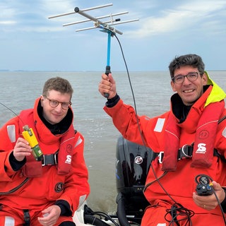 Reporter Robert und der Unterwasserlärm-Forscher Joseph Schnitzler sitzen in einem Boot. Robert hält einen Sender in der Hand und Jeff eine Antenne.