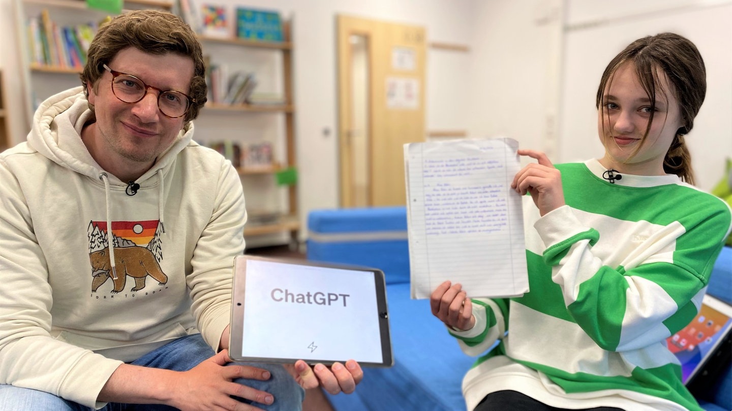 Reporter Robert und Schülerin Frida sitzen in der Schule. Robert hält ein iPad hoch, auf dem das Wort 