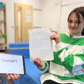 Reporter Robert und Schülerin Frida sitzen in der Schule. Robert hält ein iPad hoch, auf dem das Wort "ChatGPT" zu sehen ist. Frida hält ein Heft mit einem handgeschriebenen Aufsatz in der Hand. 