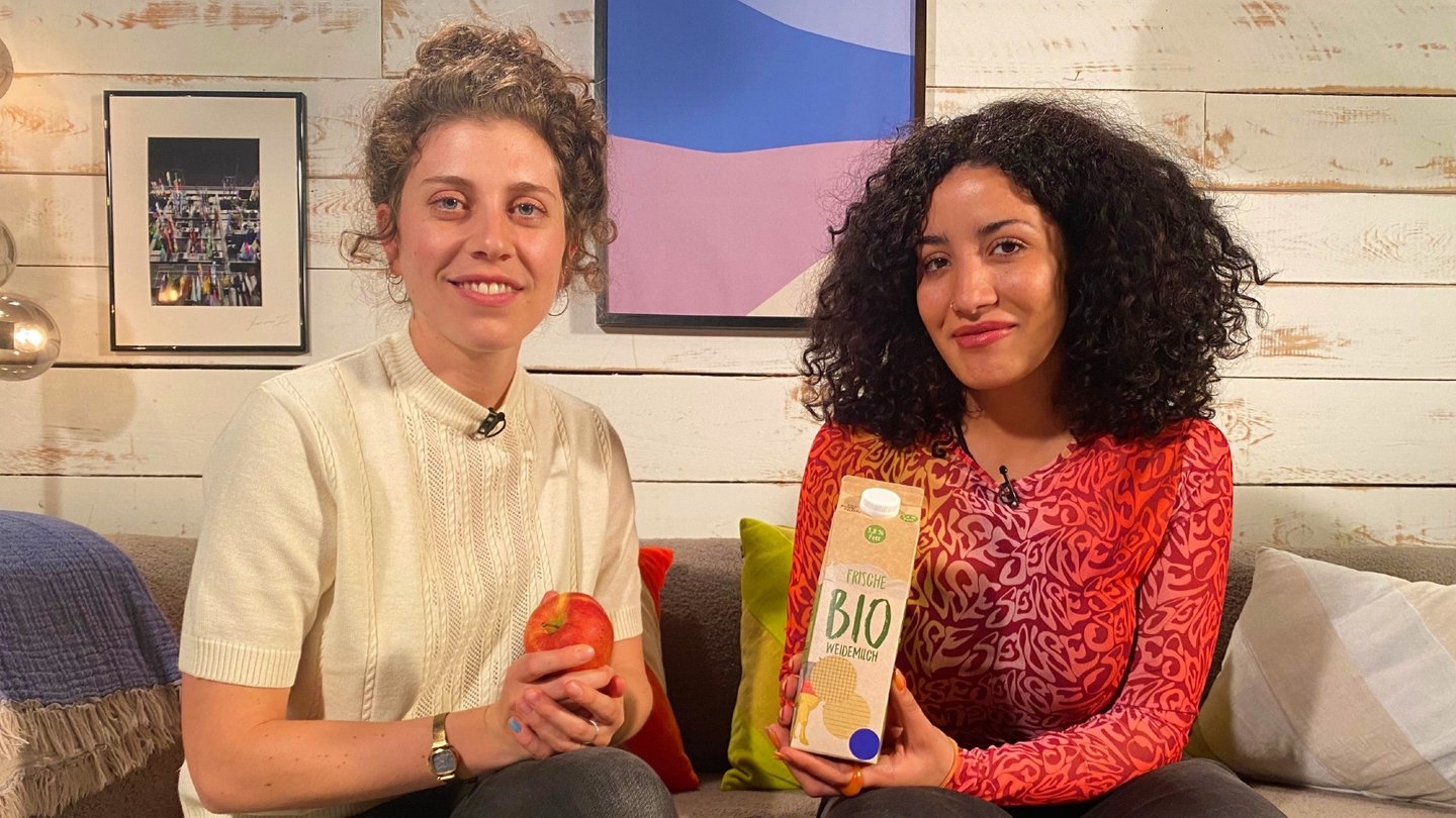 Reporterin Tessniem sitzt neben Elena Schägg auf einem Sofa. Elena hält einen Apfel in der Hand, Tessniem eine Packung Milch.