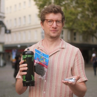 Reporter Robert Meyer hält kleine Sahnekartuschen, schwarze Luftballons und eine große Lachgas-Kartusche in der Hand.