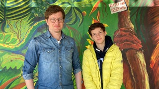 Reporter Robert und Jannis stehen vor einer bunt angemalten Wand in Jannis' Schule.