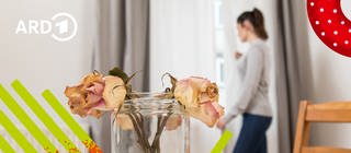 Zwei verwelkte Rosen in einer Vase auf einem Esstisch und eine Frau im Hintergrund, die traurig aus dem Fenster sieht.