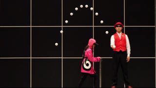 Zwei Personen stehen vor einer schwarzen Wand. Eine Person wirft einen Ball in die Höhe, seine Flugbahn ist eingezeichnet.