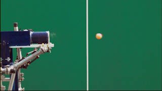Eine Ballwurfmaschine wirft einen Ball vor einer grünen Wand ab.