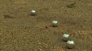 Vier silberne Boulekugeln und eine kleiner roter Ball liegen auf sandigem Boden. 