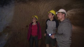 Drei Personen sind in einer Höhle und tragen Helme und eine Taschenlampe.