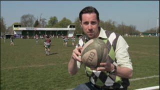 Ein Mann steht vor einem Sportplatz und hält einen Rugbyball in der Hand. Hinter ihm spielen zwei Mannschaften gegeneinander Rugby.