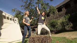 Ein Mann und eine Frau stehen vor einem Baum. Der Mann zieht ein Schwert aus einem Heuballen.