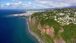 Die Küste einer Insel. Vorne blaues Meer und hohe, grüne Klippen, weiter hinten eine Stadt mit hellen Häusern.