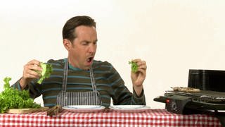 Ein Mann sitzt an einem Tisch. Er macht Salat und brät Würstchen. 